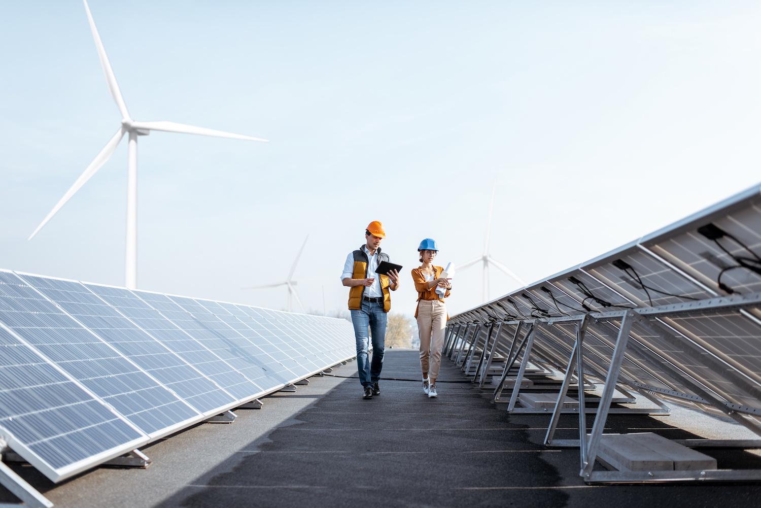 Workers walking through a solar power plant with wind turbines in the background – Achieving the SDGs with renewable energy