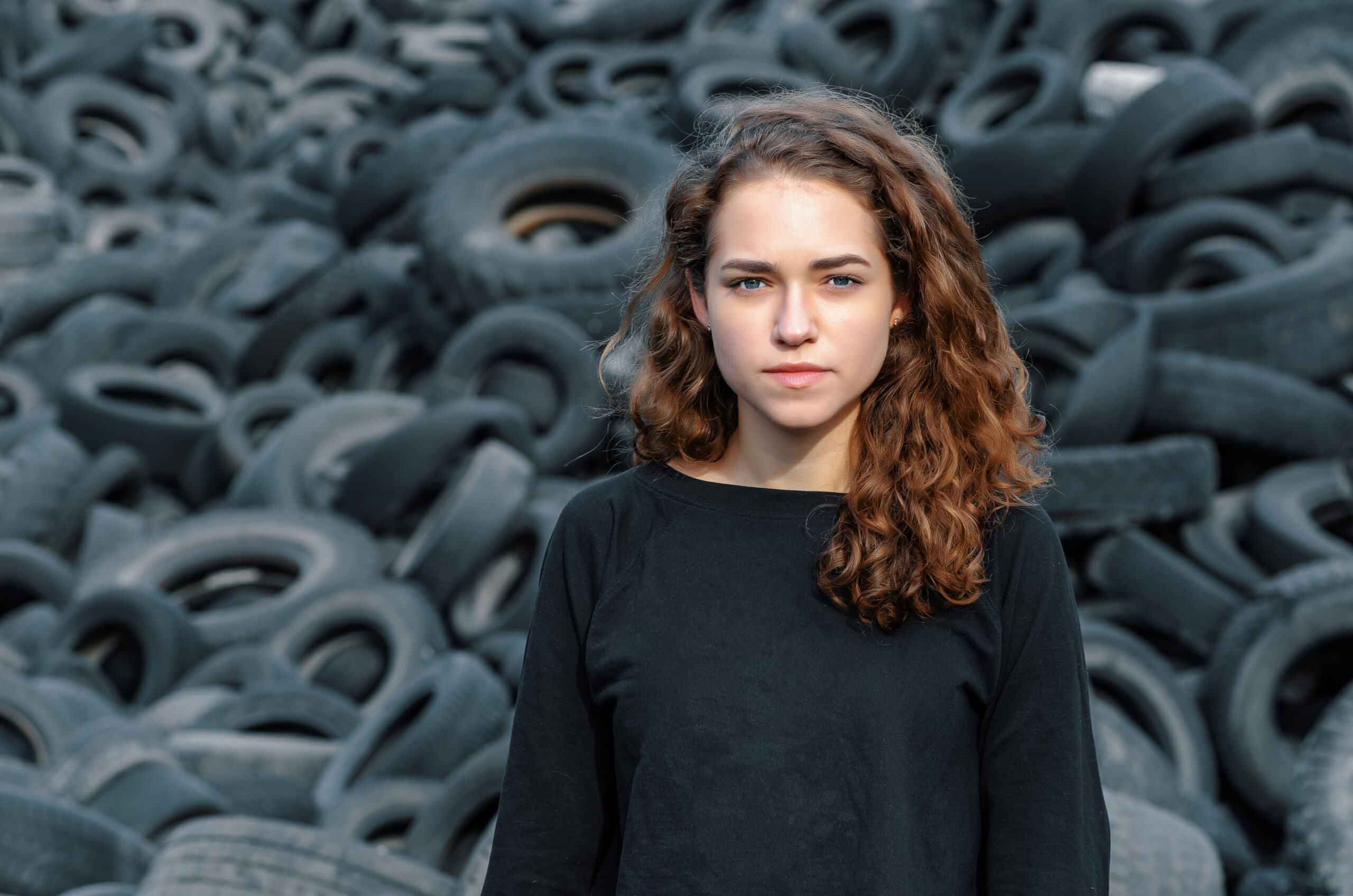 Beautiful girl, curly hair, portrait on the background of a dump of old car tires. Looking at camera.