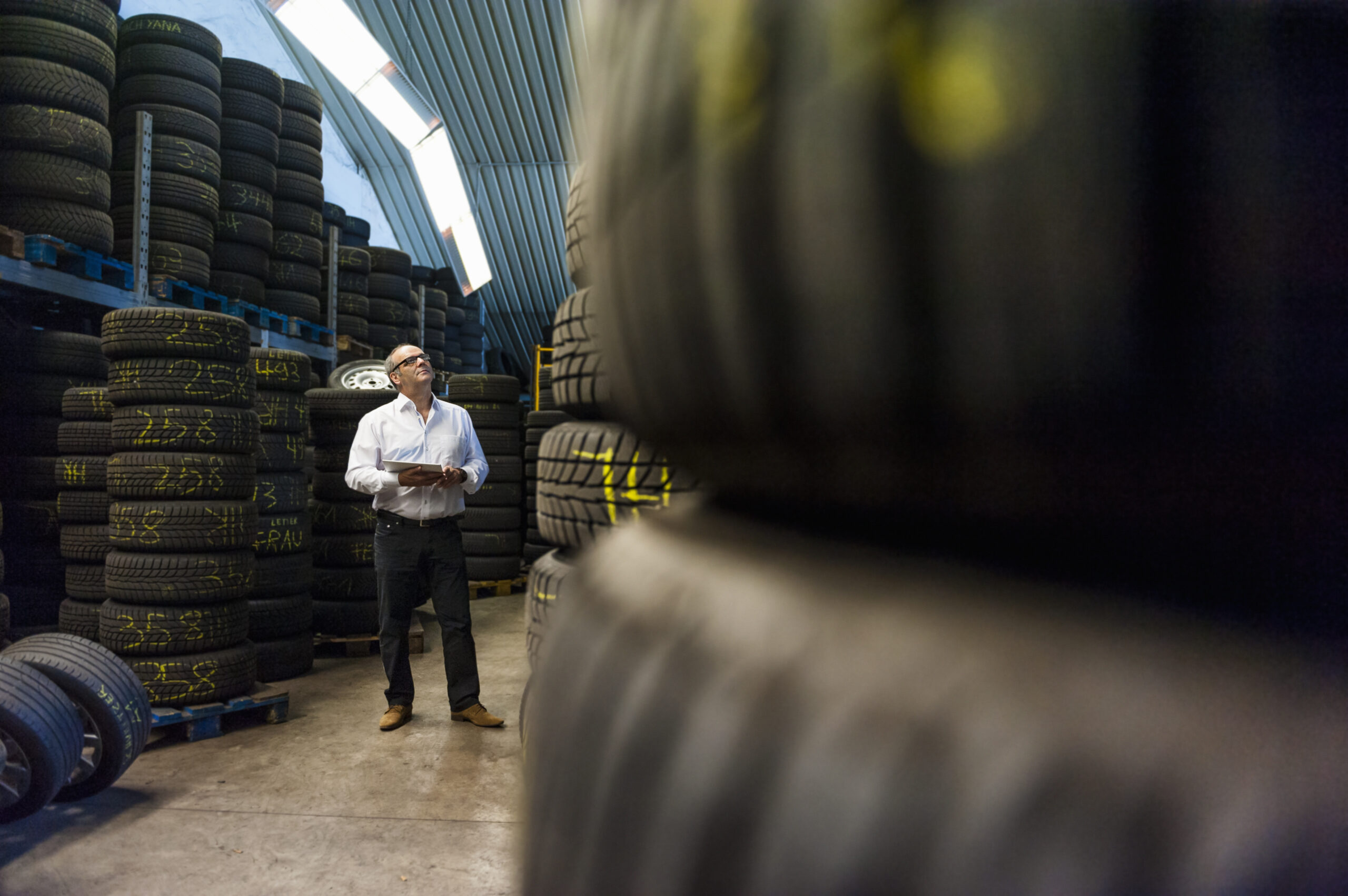 Senior businessman looking up while standing with digital tablet amidst tires